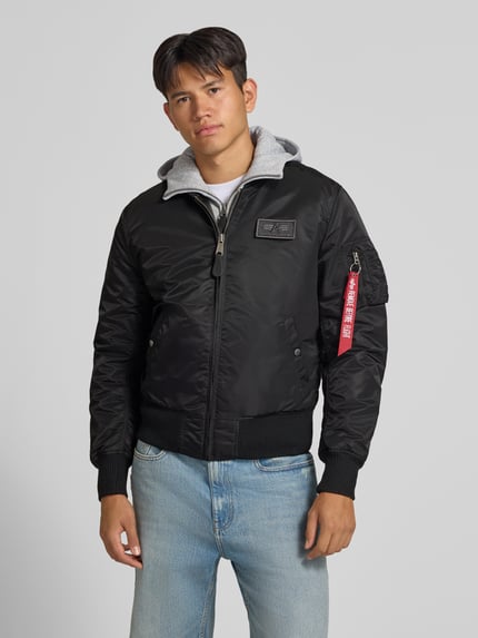 Männermodel trägt Jacke mit Reißverschlusstasche am Ärmel von Alpha Industries in Schwarz mit grauem Kapuzeinsatz, Nahaufnahme Oberkörper. Regular Fit, gerippter Stehkragen, Label-Patch, Eingrifftaschen mit Druckknopfverschluss, Brustweite 54 cm, Ärmellänge 72 cm.