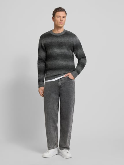 Männermodel trägt Strickpullover mit Rundhalsausschnitt von Jack & Jones Premium im Material-Mix mit Woll-Anteil, lockerer Schnitt, graue Farbverläufe, gerippte Abschlüsse, Rückenlänge und Ärmellänge 68 cm bei Größe M, Ganzkörperansicht von vorne.