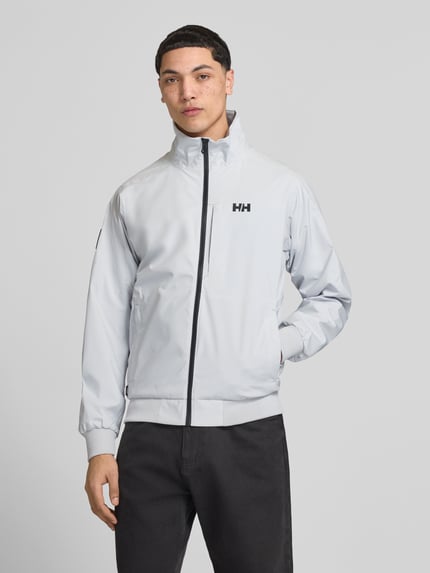 Mannenmodel draagt bomberjack met opstaande kraag model 'RACING' van Helly Hansen, recht model, ritssluiting, logoprint, zij-steekzakken, ribboorden, lichte kleur, glad oppervlak, vooraanzicht busteshot.