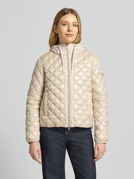 Frauenmodel trägt Regular Fit Jacke in Stepp-Optik Modell 'YOSIE' von Frieda & Freddies in Beige mit Kapuze, Reißverschluss, Eingrifftaschen, Logo-Applikation, elastische Ärmelabschlüsse, Ganzkörperansicht von vorne.