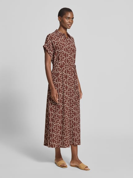 Männermodel trägt Midikleid aus wiskozy mit wiązany pasek von Gant, taliowany fason, kołnierzyk koszulowy, listwa guzikowa, długość tyłu w rozmiarze 36: 130 cm, szerokość klatki piersiowej w rozmiarze 36: 53 cm, długość rękawów w rozmiarze 36: 10 cm, wzór geometryczny w odcieniach brązu i beżu, faktura lekko zwiewna, Ganzkörperansicht von vorne.