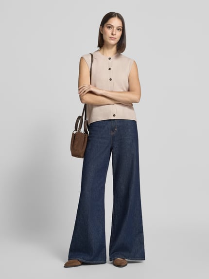 Frauenmodel trägt Regular Fit Strickweste aus reiner Schurwolle von Marc O'Polo in Beige mit dunklen Knöpfen, weite Jeans, Ganzkörperansicht von vorne. Damen Strickweste von Marc O'Polo.