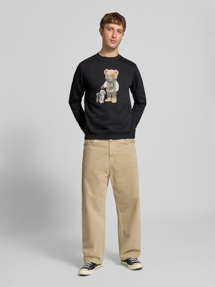 Męski model nosi czarną bluzę Sweatshirt mit Motiv-Print od CHILL PILL, mieszanka bawełny, krój regular fit, okrągły dekolt, długość tyłu w rozmiarze M: 68 cm, długość rękawów w rozmiarze M: 74 cm, nadruk z motywem, widoczna cała sylwetka z przodu.