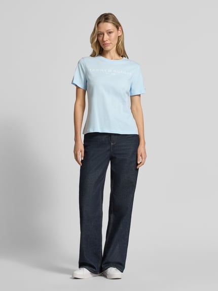 Frauenmodel trägt Regular Fit T-Shirt aus reiner Baumwolle von Tommy Hilfiger in Hellblau, unifarben, Logo-Schriftzug, kurze Ärmel, Rundhals, Jersey, Ganzkörperansicht von vorne.