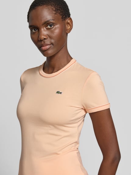 Frauenmodel trägt Slim Fit T-Shirt aus Baumwoll-Mix von Lacoste in Apricot mit Logo-Stitching und Kontrastpaspel, Nahaufnahme Oberkörper.