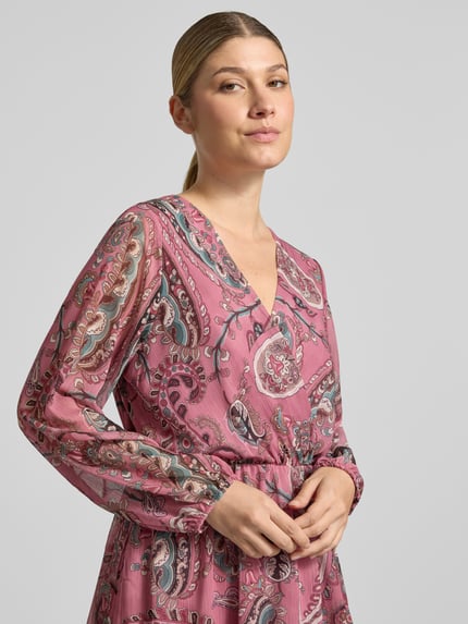 Kobiece modelka ma na sobie sukienkę ZABAIONE Knielanges Kleid mit V-Ausschnitt Modell 'Lu44ciaa', wzór na całej powierzchni, kopertowy styl, dekolt w serek, taliowany krój, długość do kolan, długość tyłu w rozmiarze S: 92 cm, szerokość w rozmiarze S: 35 cm, długość rękawów w rozmiarze S: 28 cm, różowe i szare wzory paisley, faktura lekko marszczona, zbliżenie na górną część ciała, perspektywa z przodu.