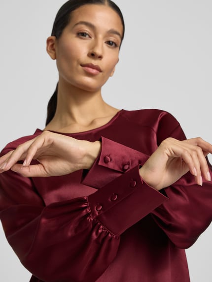 Modelka kobieca nosi knielanges Cocktailkleid mit Rundhalsausschnitt Modell 'Aurea' von GUIDO MARIA KRETSCHMER WOMAN, bordowy materiał z połyskiem, długi rękaw z mankietami, materiał z domieszką stretchu, prosty krój, ujęcie w zbliżeniu na górną część ciała.