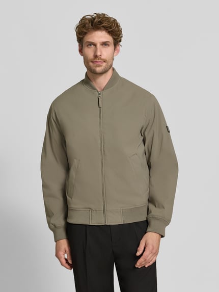 Męski model nosi bomberkę z zamkiem błyskawicznym i wpuszczanymi kieszeniami Bomberjacke mit Reißverschluss und Eingrifftaschen von JACK & JONES premium, krój regular fit, stójka, długość tyłu w rozmiarze M: 66 cm, szerokość ramion w rozmiarze M: 42 cm, szerokość klatki piersiowej w rozmiarze M: 60 cm, długość rękawów w rozmiarze M: 68 cm, jednolity beżowy kolor, prosta faktura, całkowity widok z przodu.