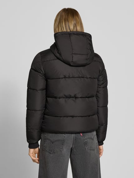 Frauenmodel trägt schwarze Steppjacke mit Kapuze von PIECES, klare Steppnähte, glatte Textur, Ganzkörperansicht Rückansicht.