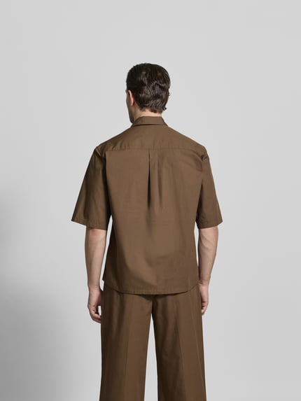 Männermodel trägt Relaxed Fit Freizeithemd mit Kentkragen von Officine Générale in Braun, reine Baumwolle, Ganzkörperansicht Rückansicht. Rückenlänge bei Größe M: 74 cm, Schulterbreite: 60 cm, Ärmellänge: 33 cm, Knopfleiste, Halbarm.