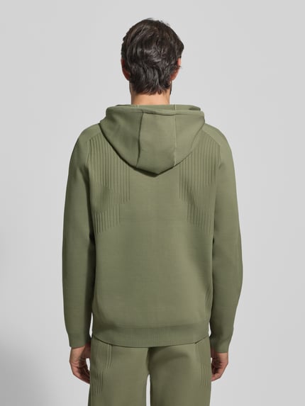Męski model nosi kurtkę treningową Regular Fit Trainingsjacke mit Jacquard Elementen Modell 'ACTIVE PUSH' od BOSS Green, oliwkowy kolor, żakard, faktura, kaptur z tunelem, długie rękawy, wykończenia prążkowane, kieszenie na zamek, widok całej sylwetki od tyłu.