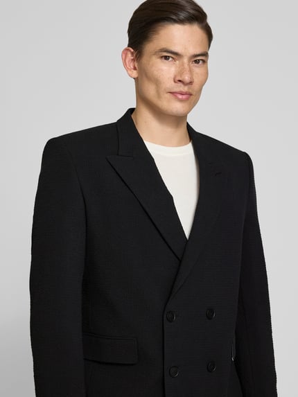 Męski model nosi Slim Fit Anzugsakko aus Baumwoll-Mix Modell 'HENRY' od HUGO, czarna gładka faktura, krój slim fit, detal: krótka dwurzędowa listwa guzikowa, klasyczny kołnierz z klapami, kieszenie z patkami; materiał: bawełniany mix; zbliżenie górnej części ciała z przodu.