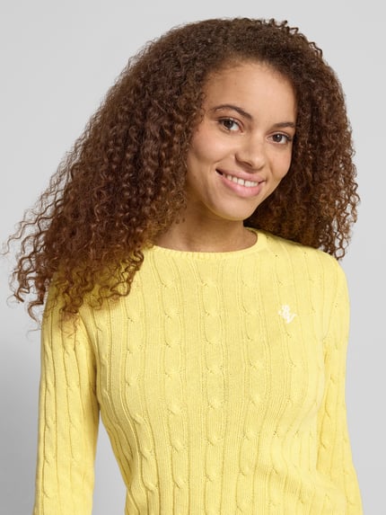 Kobiece modelka nosi sweter z dzianiny 'Strickpullover mit Zopfmuster und Label-Stitching' od REVIEW, mieszanka bawełny, krój regular fit, ścieg warkoczowy, okrągły dekolt, wyhaftowane logo, długość tyłu w rozmiarze S: 52 cm, długość rękawa w rozmiarze S: 69 cm, jasny żółty kolor, tekstura dzianinowa, widoczny ścieg warkoczowy, całkowite ujęcie górnej części ciała z przodu.