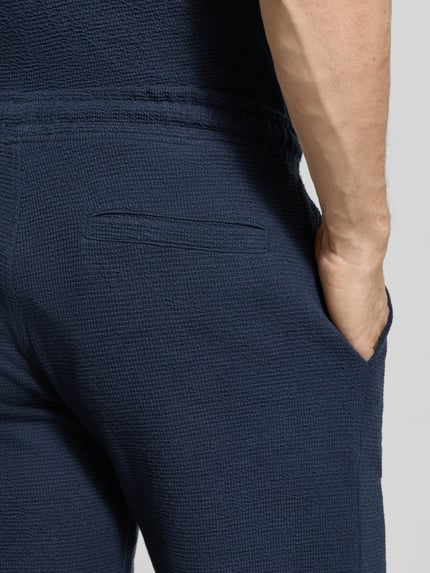 Männermodel trägt Sweatshorts mit elastischem Bund von MCNEAL in Blau mit Strukturmuster, Gesäßtasche, Elastischer-Material-Mix, Nahaufnahme Rückansicht.