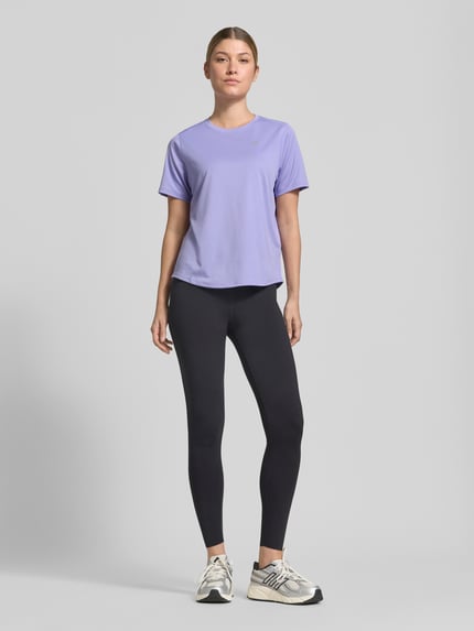 Frauenmodel trägt T-Shirt mit Rundhalsausschnitt und Logo-Print von ASICS in Flieder, kombiniert mit schwarzen Leggings, Ganzkörperansicht von vorne. Damen T-Shirt, gerader Schnitt, leicht abgerundeter Saum, Rückenlänge bei Größe S 60 cm.