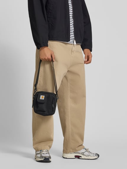 Männermodel trägt zwarte Crossbody Bag met logopatch van Carhartt Work In Progress, verstelbare schouderband, hoofdvak met ritssluiting, voorvak, binnenzak met rits, afneembare schouderband, breedte 17 cm, lengte 18 cm, diepte 6 cm, bandlengte 80 cm, ganzkörperansicht van voren.