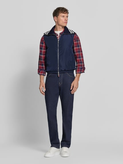 Männermodel trägt Weste mit Kontraststreifen von Dsquared2, navyblau mit weißen Akzenten, Rippkragen, Zweiwege-Reißverschluss, elastischer Bund, locker geschnitten, kombiniert mit kariertem Hemd und Jeans, Ganzkörperansicht von vorne.