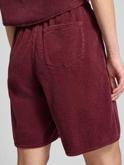 Männermodel trägt Shorts mit elastischem Bund Modell 'PADOW' von American Vintage, bordowy kolor, sztruksowa faktura, luźny krój, kieszeń, czysta wiskoza, Szerokość w rozmiarze S: 37 cm, długość wewnętrzna nogawki S: 20 cm, zdjęcie tyłu w ujęciu całej sylwetki.