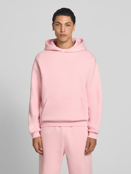 Männermodel trägt Hoodie mit Logo-Print von REVIEW im Baumwoll-Mix, lockere Passform, Kängurutasche, rosa, Ganzkörperansicht von vorne.