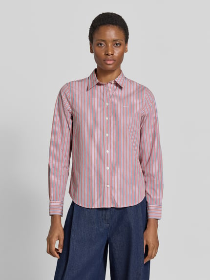 Frauenmodel trägt Regular Fit Langarmbluse aus reiner Baumwolle von GANT, rot-blau-weiß gestreift, kombiniert mit blauer Hose, Ganzkörperansicht von vorne.