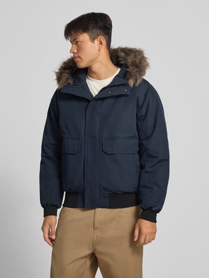 Männermodel trägt dunkelblaue Bomberjacke mit Label-Patch Modell 'Everest' von Superdry, Kunstfellkapuze, Pattentaschen, Ganzkörperansicht von vorne. Herren Bomberjacke, Regular Fit, Reiß- und Druckknopfverschluss.