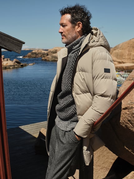 Männermodel trägt Mantel mit Kapuze und Label-Patch von NAUTGÅRD in Beige, kombiniert mit Strickpullover, Ganzkörperansicht von vorne. Herren Jacke, gerader Schnitt, Zweiwege-Reißverschluss, seitliche Reißverschlusstaschen, gestrickte Ärmelabschlüsse, Größe M: Rückenlänge 100 cm, Schulterbreite 52 cm, Brustweite 60 cm, Ärmellänge 65 cm.
