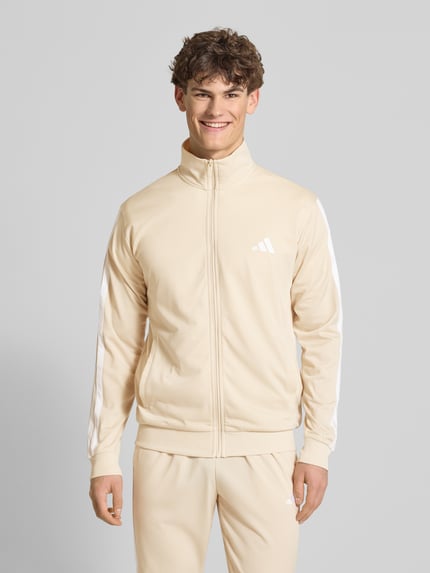 Männermodel trägt Regular Fit Sweat-Anzug mit Kontraststreifen von adidas Sportswear in Beige mit weißen Streifen, unifarben, Ganzkörperansicht von vorne.