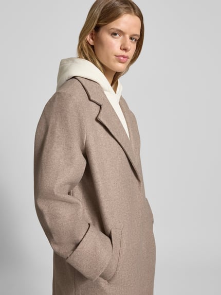 Kobiece model prezentuje płaszcz z klapami Mantel mit Reverskragen Modell 'CADDY' od Noisy May, beżowy, tekstura miękka, widok z przodu, ujęcie górnej części sylwetki. Relaxed Fit, kieszenie na ręce, mankiety z wywinięciem, długość tyłu 120 cm, szerokość ramion 43 cm, szerokość klatki 52 cm, długość rękawa 65 cm.