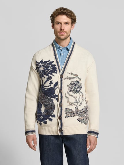 Männermodel trägt Regular Fit Strickjacke mit V-Ausschnitt Modell 'INTARSIA' von Scotch & Soda, Allover-Muster, Creme und Blau, Ganzkörperansicht von vorne. Baumwoll-Mix mit Woll-Anteil, Knopfleiste, Kontext: Rückenlänge 71 cm, Ärmellänge 60 cm.