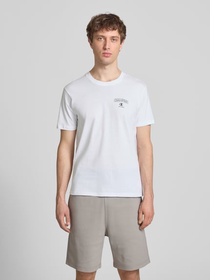 Männermodel trägt T-Shirt mit Logo und Rundhalsausschnitt von CHAMPION in Weiß, reine Baumwolle, Regular Fit, Ganzkörperansicht von vorne.