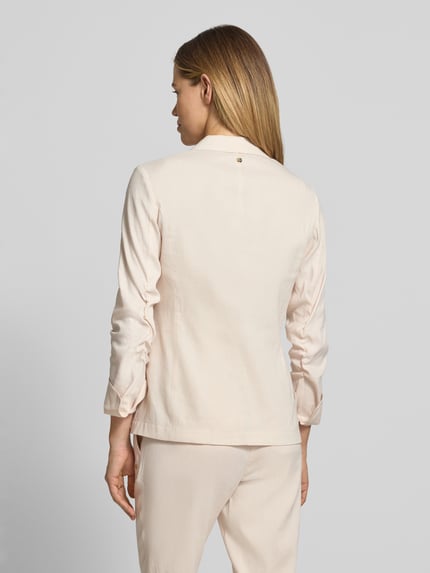 Modelka nosi Blazer aus Leinen-Mix mit Reverskragen von Marc Cain, mieszanka lnu i wiskozy, taliowany, kołnierz z połami, zapięcie na guzik, kieszenie z patką, rękawy z zakładką, długość tyłu w rozmiarze 36: 62 cm, szerokość w rozmiarze 36: 44 cm, długość rękawów w rozmiarze 36: 52 cm, jasny kolor, gładka tekstura, widok całej sylwetki z tyłu.