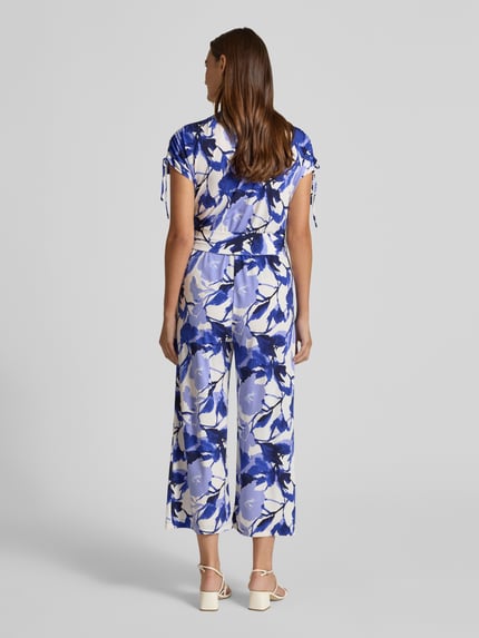 Kobiecy model nosi Relaxed Fit Jumpsuit mit V-Ausschnitt od Betty Barclay, elastyczna mieszanka materiałów, taliowany krój, dekolt w serek, krótki rękaw, kwiatowy nadruk na całej powierzchni, wpuszczane kieszenie, odcienie niebieskiego i bieli, całkowita długość 132 cm, długość tyłu 43 cm, szerokość w rozmiarze 36: 49 cm, długość rękawów 22 cm, długość wewnętrzna nogawki 61 cm, widok całej sylwetki z tyłu.