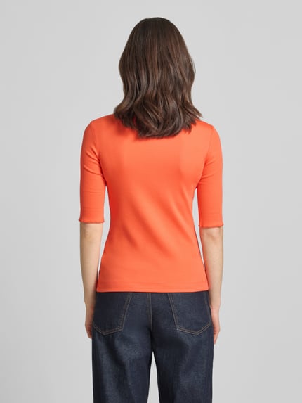 Frauenmodel trägt T-Shirt mit Label-Applikation von Marc Cain in Orange, körpernaher Schnitt, U-Ausschnitt, mitteldicke Baumwoll-Mix-Textur, Ganzkörperansicht Rückansicht.