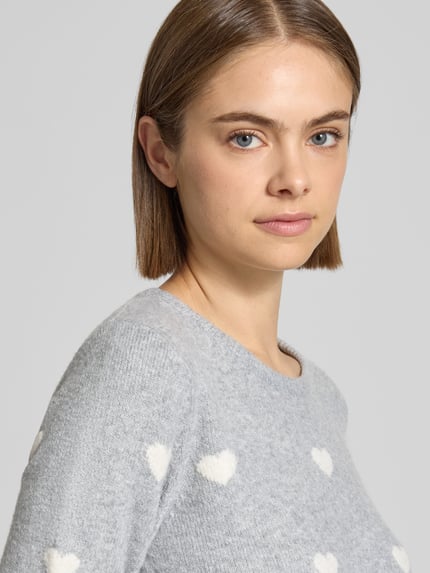 Frauenmodel trägt Strickpullover mit geripptem Saum von REVIEW, grau mit weißen Herzmustern, weiche Textur, Nahaufnahme Oberkörper.