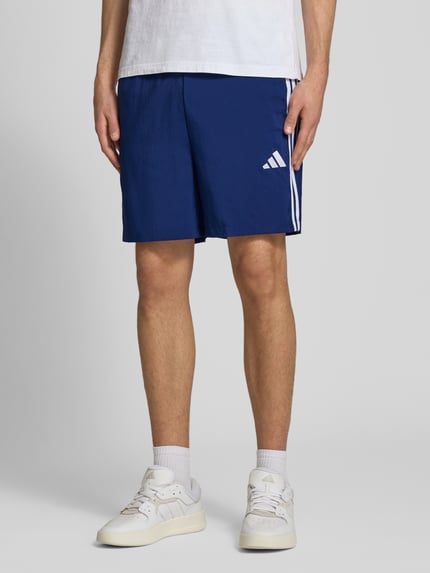 Männermodel trägt Regular Fit Sport-Shorts mit elastischem Bund von adidas Sportswear, unifarben blau, Kontraststreifen, Nahaufnahme Oberschenkel.