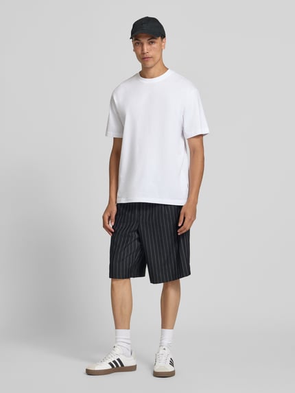 Męski model nosi szorty z elastycznym pasem model 'CRISTIANO' od Redefined Rebel, mieszanka materiałów z dodatkiem streczu, krój relaxed fit, szerokość w rozmiarze M: 42 cm, długość wewnętrzna nogawki: 27 cm, ciemne kolory z białymi paskami, gładka faktura, całopostaciowy widok z przodu.