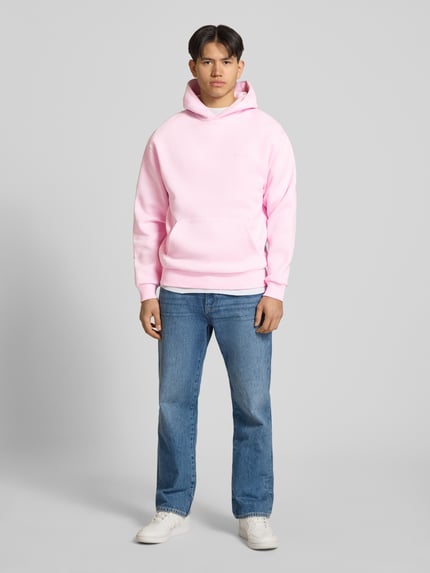 Męskimodel nosi Hoodie mit Känguru-Tasche von REVIEW, różowa mieszanka bawełny, luźny krój, kieszeń kangurka, detal z logo, elastyczne wykończenia, długość tyłu w rozmiarze m: 68 cm, długość rękawów w rozmiarze m: 62 cm, całopostaciowy widok z przodu.