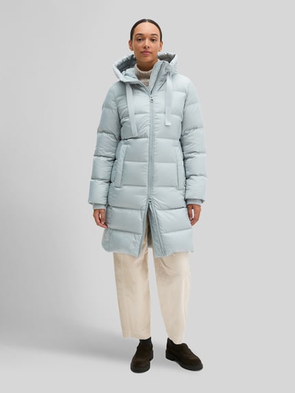 Frauenmodel trägt Regular Fit Daunenmantel mit Label-Print von Marc O'Polo in Hellblau, matte Oberfläche, Ganzkörperansicht von vorne.