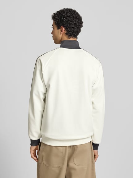 Męski model nosi Regular Fit Trainingsjacke mit Vereins Logo Applikation Modell 'BLACAS' od adidas Originals, krój regular fit, gładki kremowy materiał z czarnym kołnierzem i ściągaczami, widoczne detale treningowej bluzy, widok całej sylwetki z tyłu