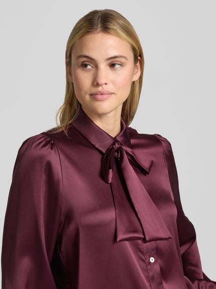 Vrouwenmodel draagt Regular Fit Hemdbluse aus Satin van Christian Berg Woman Selection in bordeauxrood met sierstrik, satijnen textuur, blousekraag, lange mouwen, detailopname bovenlichaam.