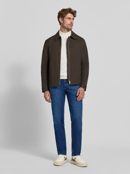 Männermodel trägt blaue Straight Fit Jeans aus Baumwoll-Mix Modell 'Cadiz' von Brax, kombiniert mit weißem Pullover und brauner Jacke, Ganzkörperansicht von vorne, Herren Straight Fit Jeans von Brax
