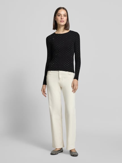 Modelka kobieca nosi Langarmshirt mit schimmernden Details od VERO MODA, czarny w białe kropki, długi rękaw, dekolt okrągły, błyszcząca tekstura, widok na całą sylwetkę z przodu.