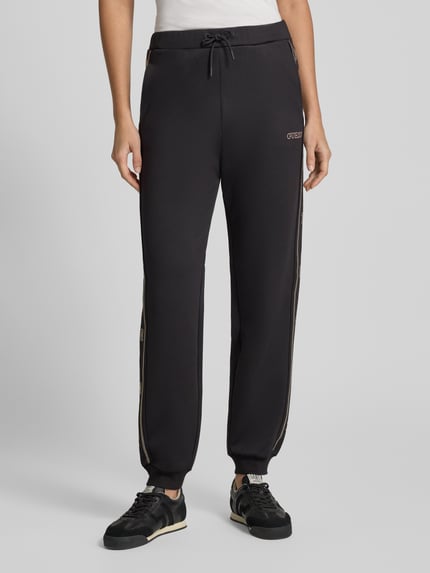 Kobieta nosi czarne spodnie dresowe Sweatpants mit Label-Detail Modell 'OCTAVIA' od GUESS Activewear, krój regular fit, elastyczny pas, sznurek, miękki materiał z dodatkiem wiskozy, długość nogawki 73 cm w rozmiarze S, widok całej sylwetki z przodu.