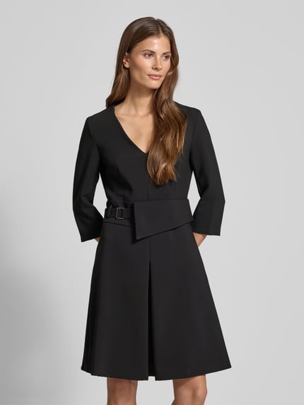Frauenmodel trägt knielanges Cocktailkleid mit V-Ausschnitt von Rinascimento in Schwarz, Material-Mix, tailliert, 3/4-Ärmel, Taillengürtel, Ganzkörperansicht von vorne.