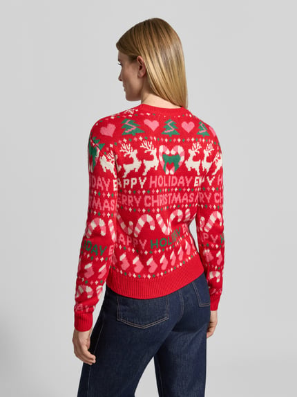 Frauenmodel trägt Pullover mit Weihnachtsmotiv-Frontprint von VERO MODA in mehrfarbiger Strickoptik, grafischer Motiv-Print, Rippbündchen, Ganzkörperansicht Rückansicht.