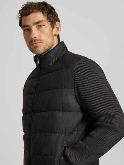 Männermodel trägt Steppjacke mit Stehkragen und Zweiwege-Reißverschluss von Christian Berg Men in Schwarz, Nahaufnahme Oberkörper, Regular Fit, feine Rippenstruktur an den Ärmeln, strukturierte Oberfläche, Reißverschlusstaschen, KI-modifiziertes Bild.