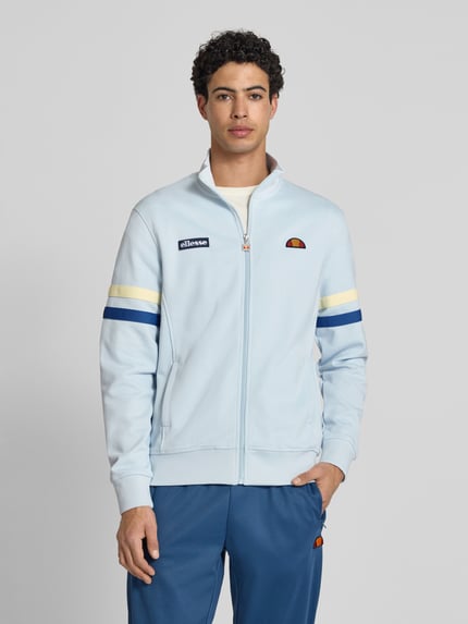 Männermodel trägt Sweatjacke aus Baumwoll-Mix Modell 'ROMA' von Ellesse in Hellblau mit Label-Details, Stehkragen, Eingrifftaschen, Gerade Passform, elastische Abschlüsse, durchgehender Reißverschluss; Rückenlänge 70 cm, Ärmellänge 68 cm, Ganzkörperansicht von vorne.