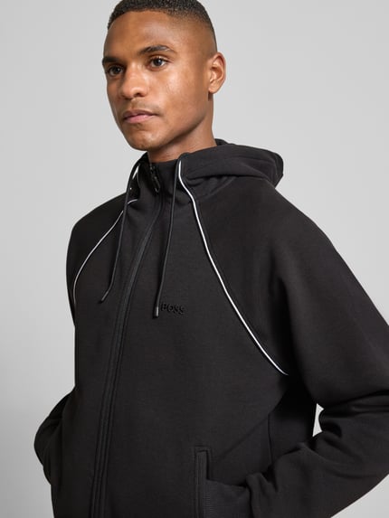 Männermodel trägt Regular Fit Sweatjacke aus Baumwoll-Mix von BOSS Green in Schwarz mit Kontraststreifen und Kapuze, Nahaufnahme Oberkörper.