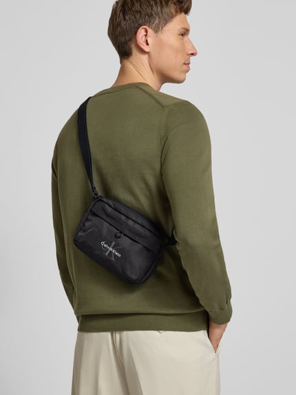 Mannelijk model draagt Crossbody Bag met Label-Stitching van CK Calvin Klein, hoofvak met ritssluiting, verstelbare schouderband, breedte 21 cm, lengte 15 cm, diepte 6 cm, bandlengte 80 cm, schoudertas, geheel van achteren bekeken.
