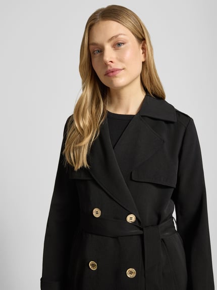 Kobieta nosi dwurzędowy trencz z wiązanym paskiem MICHAEL Michael Kors, krój regular fit, kołnierz z połami, zbliżenie na górę. Długość tyłu w rozmiarze S: 47 cm, szerokość ramion w rozmiarze S: 40 cm, szerokość klatki piersiowej w rozmiarze S: 50 cm, długość rękawów w rozmiarze S: 63 cm.
